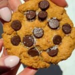 A close-up of hands holding a chocolate chip cookie with visible sea salt flakes on top.