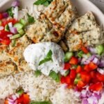 A bowl of white rice topped with grilled kebab pieces, diced red onion, red and green bell peppers, and a dollop of yogurt garnished with a mint leaf.