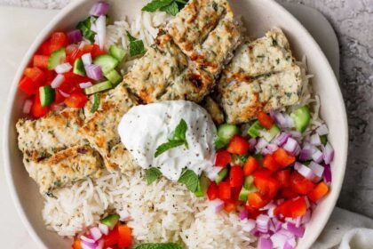 A bowl of white rice topped with grilled kebab pieces, diced red onion, red and green bell peppers, and a dollop of yogurt garnished with a mint leaf.