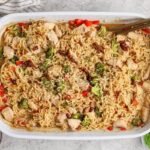 Rectangular white baking dish filled with cooked ramen noodles, chicken pieces, broccoli, and red peppers, garnished with sesame seeds and sauce, with chopsticks resting on the dish.