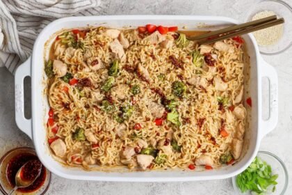 Rectangular white baking dish filled with cooked ramen noodles, chicken pieces, broccoli, and red peppers, garnished with sesame seeds and sauce, with chopsticks resting on the dish.