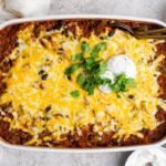 A baked casserole topped with melted cheese, black beans, sour cream, and chopped cilantro, surrounded by bowls of lime, shredded cheese, and extra sour cream.