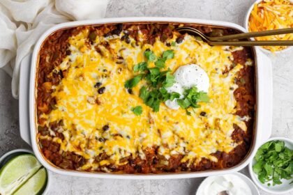 A baked casserole topped with melted cheese, black beans, sour cream, and chopped cilantro, surrounded by bowls of lime, shredded cheese, and extra sour cream.