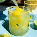A glass decorated with small pineapples is filled with yellow frozen drink and ice, with juice being poured in. Lime slices are on the blue surface beside the glass.