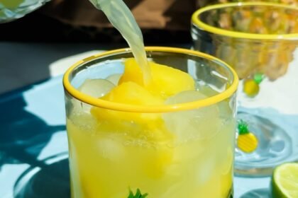 A glass decorated with small pineapples is filled with yellow frozen drink and ice, with juice being poured in. Lime slices are on the blue surface beside the glass.