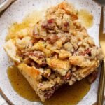 A close-up of a slice of pecan streusel bread pudding topped with pecans and syrup, served on a speckled plate with a fork.