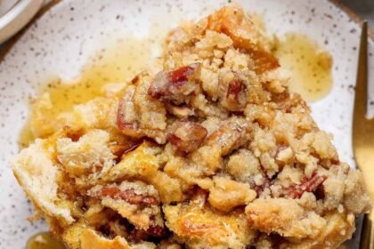 A close-up of a slice of pecan streusel bread pudding topped with pecans and syrup, served on a speckled plate with a fork.