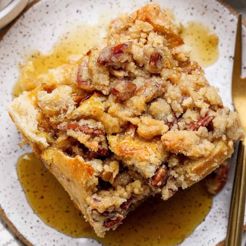 A close-up of a slice of pecan streusel bread pudding topped with pecans and syrup, served on a speckled plate with a fork.