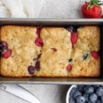 A rectangular loaf pan contains a baked berry bread with visible raspberries and blueberries. Fresh strawberries, blueberries, and raspberries are placed around the pan.