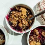 Three small white ramekins filled with baked berry dessert, topped with peanut butter and chocolate chips, with metal spoons and a red-striped cloth nearby.
