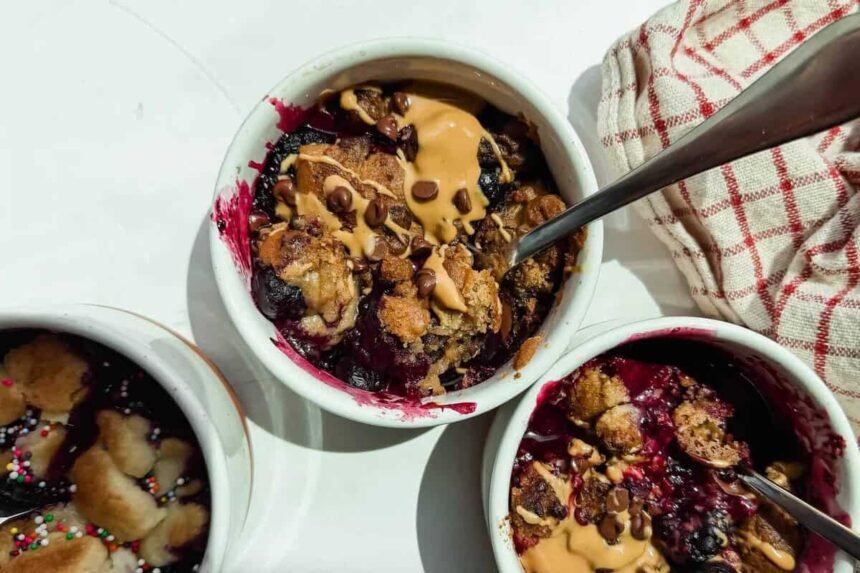 Three small white ramekins filled with baked berry dessert, topped with peanut butter and chocolate chips, with metal spoons and a red-striped cloth nearby.