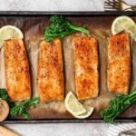 Broiled salmon fillets on a baking sheet garnished with lemon slices, broccolini, and a bowl of seasoning.