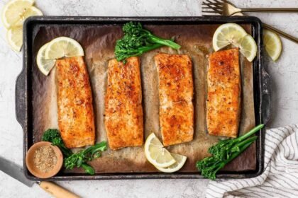 Broiled salmon fillets on a baking sheet garnished with lemon slices, broccolini, and a bowl of seasoning.