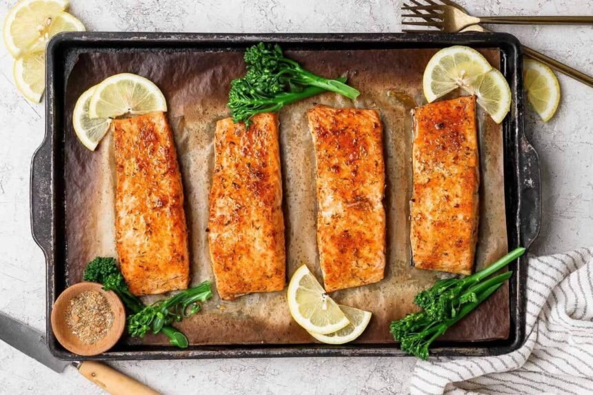 Broiled salmon fillets on a baking sheet garnished with lemon slices, broccolini, and a bowl of seasoning.