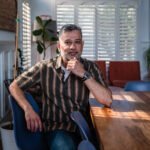 A South Asian man sits by the window at a table. He rests his elbow on the table with his hand on his chin. He looks contemplative.