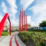 Landscape shot of ARTivity on the Green in Winston-Salem during daytime. Blue skies and greenery with flowers in bloom. Red sculptures in foreground with city in background. Spring or summer season.