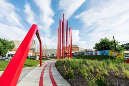Landscape shot of ARTivity on the Green in Winston-Salem during daytime. Blue skies and greenery with flowers in bloom. Red sculptures in foreground with city in background. Spring or summer season.