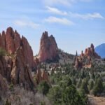 Towering red sandstone spires and cliffs surrounded by evergreens at Garden of the Gods, with dramatic peaks and blue sky.