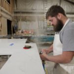 A man works with a polishing tool on the edge of a countertop. Dust can be seen flying in the air around the tool. Some of the dust has settled in the man's hair.