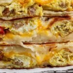 Three breakfast stuffed flatbreads stacked, filled with scrambled eggs, sausage, melted cheese, and some visible tomato pieces.