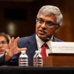 Jay Bhattacharya speaks while sitting at a table with his nameplate, and two water bottles, in front of him.
