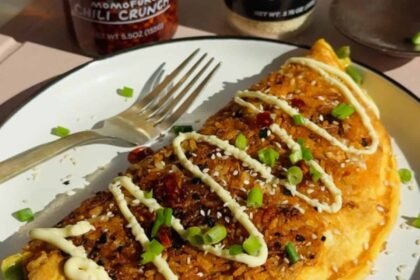 Omelette in a pan topped with chopped carrots, edamame, corn, and fresh herbs, shown in natural sunlight.
