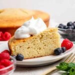 A slice of yellow cake topped with whipped cream, served on a plate with fresh raspberries and blueberries; whole cake and more berries in the background.