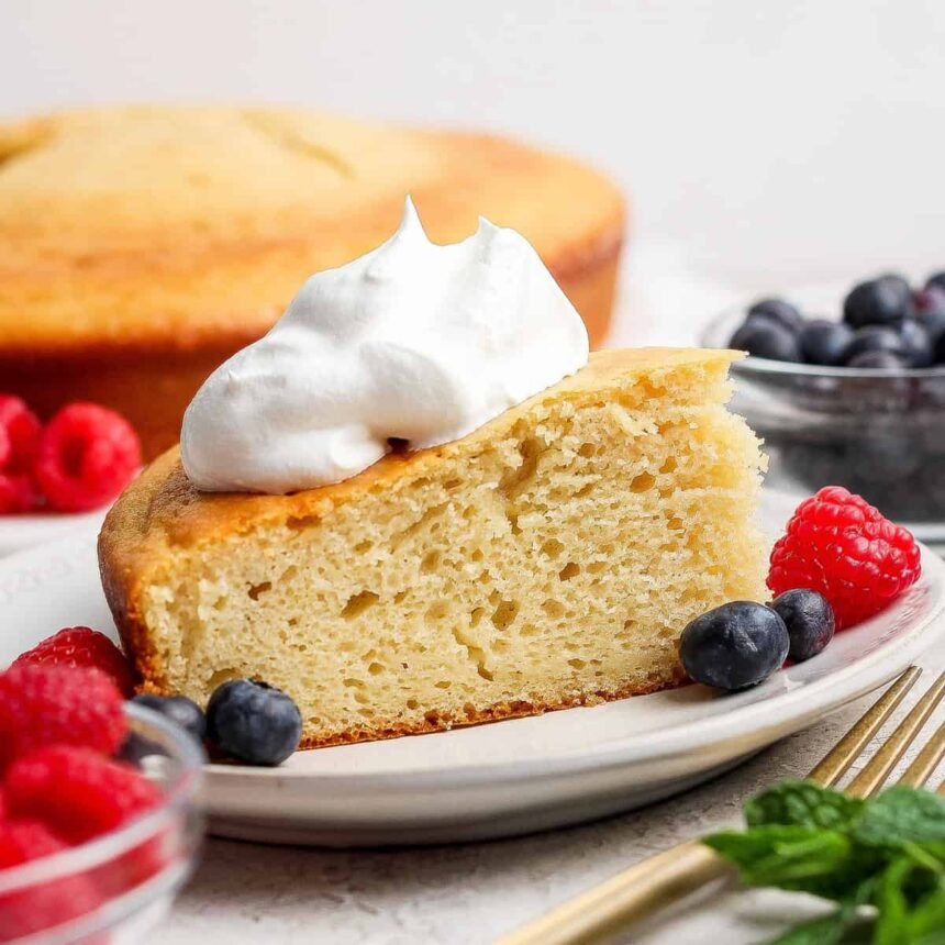 A slice of yellow cake topped with whipped cream, served on a plate with fresh raspberries and blueberries; whole cake and more berries in the background.