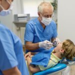 An older male dentist performs a cleaning on a young boy patient. A female hygienist is seen blurred in the foreground.