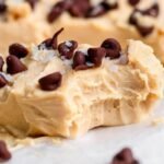 A creamy peanut butter fudge piece topped with chocolate chips and flaky salt, showing a bite taken out, resting on parchment paper.