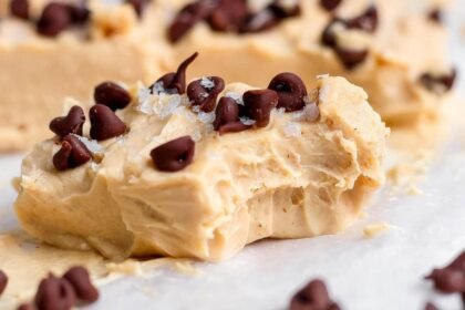 A creamy peanut butter fudge piece topped with chocolate chips and flaky salt, showing a bite taken out, resting on parchment paper.