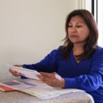 A Hispanic woman sits at a table, reading medical bills from a red file folder in front of her.