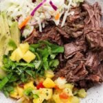 A bowl containing shredded beef, sliced avocado, coleslaw, chopped cilantro, and pineapple salsa.