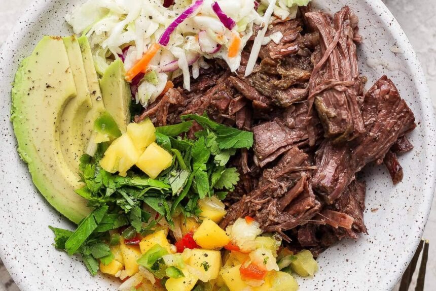 A bowl containing shredded beef, sliced avocado, coleslaw, chopped cilantro, and pineapple salsa.