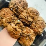 A glass dish containing seven chunky cookies drizzled with light brown icing and sprinkled with mini chocolate chips, one cookie being lifted with a pink spatula.