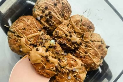 A glass dish containing seven chunky cookies drizzled with light brown icing and sprinkled with mini chocolate chips, one cookie being lifted with a pink spatula.