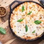 A cast iron skillet filled with baked cheesy pasta topped with melted cheese, fresh basil leaves, and sprinkled red pepper flakes. The skillet is on a wooden board with a small bowl of red pepper flakes and basil nearby.