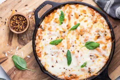A cast iron skillet filled with baked cheesy pasta topped with melted cheese, fresh basil leaves, and sprinkled red pepper flakes. The skillet is on a wooden board with a small bowl of red pepper flakes and basil nearby.