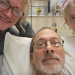 An older man lies in a hospital bed. His wife and daughter are on either side of him, smiling.