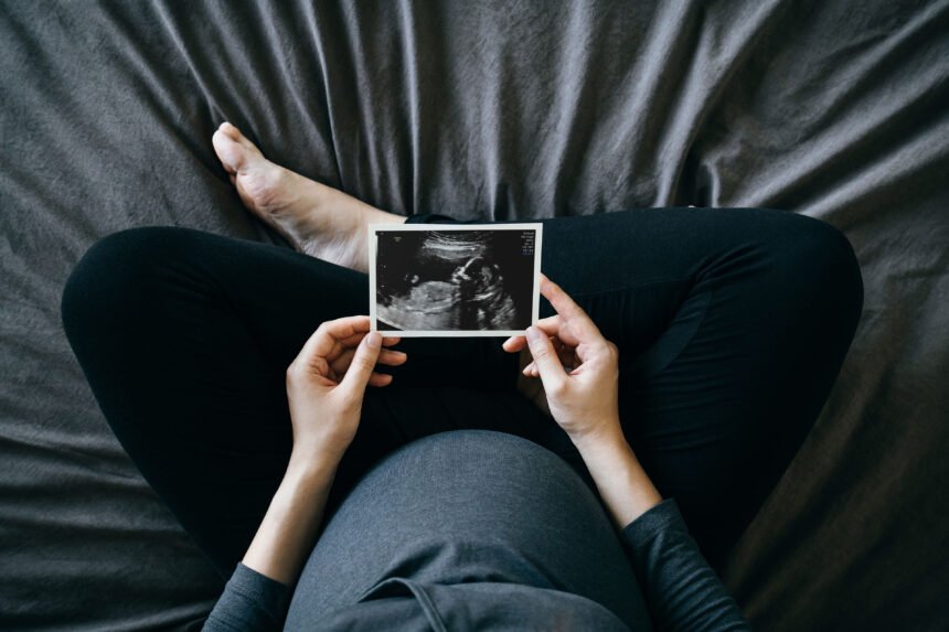 A high-angle photograph of a pregnant person sitting cross-legged on a bed and holds a sonogram of a baby.