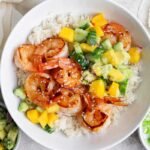 A bowl of white rice topped with grilled shrimp, diced mango, cucumber, avocado, and sprinkled sesame seeds, with side bowls of salsa and chopped green onions.