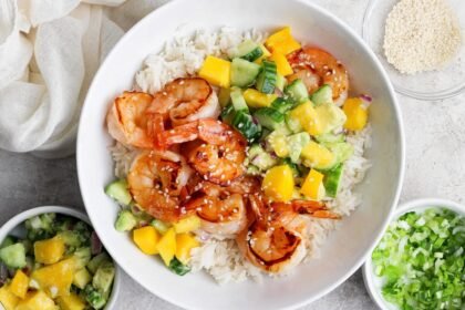 A bowl of white rice topped with grilled shrimp, diced mango, cucumber, avocado, and sprinkled sesame seeds, with side bowls of salsa and chopped green onions.