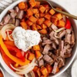 A bowl filled with diced steak, roasted sweet potatoes, saut&eacute;ed onions and bell peppers, topped with a dollop of sour cream and black pepper, with a fork resting inside.