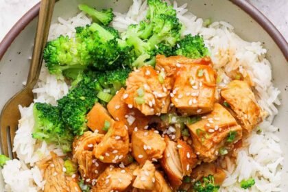 A bowl of white rice topped with cooked chicken pieces in sauce, broccoli florets, and a sprinkle of sesame seeds, with a fork on the side.