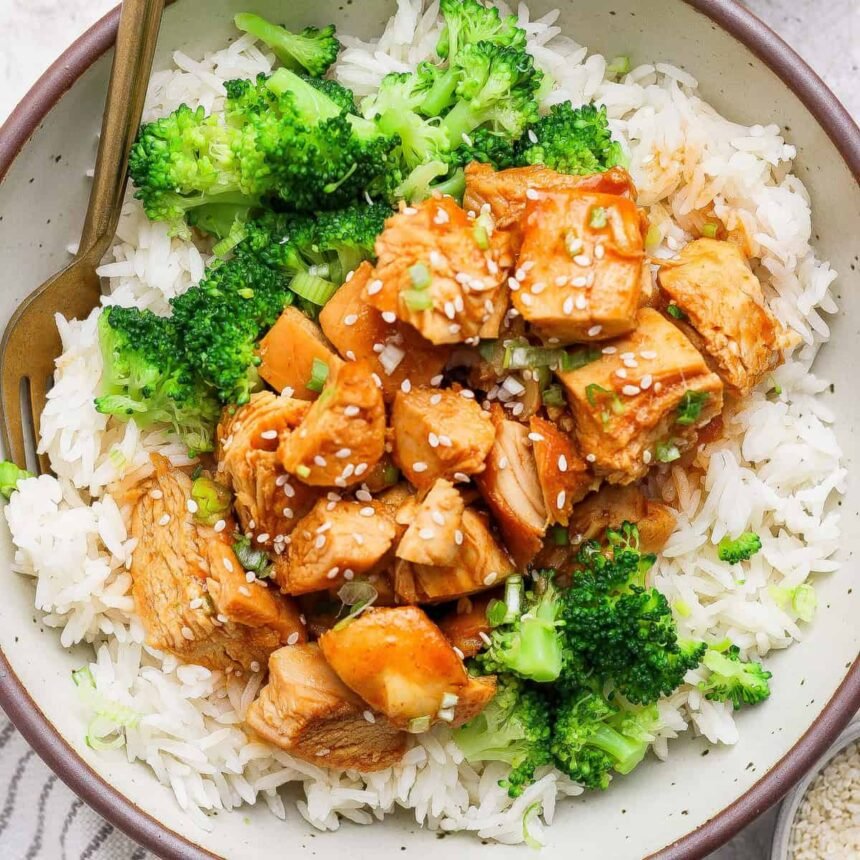 A bowl of white rice topped with cooked chicken pieces in sauce, broccoli florets, and a sprinkle of sesame seeds, with a fork on the side.