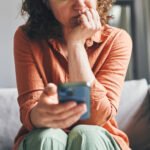A woman sitting on a couch indoors, appearing focused and concerned while looking at her smartphone.
