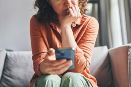 A woman sitting on a couch indoors, appearing focused and concerned while looking at her smartphone.