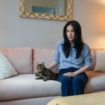 A woman sits on a couch with a tabby cat beside her. Her hand gently rests beside her cat.