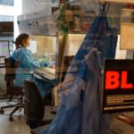 A woman in a lab gear works at a lab station surrounded by equipment. She is seen behind a glass door. A label on the glass reads "BL2+"