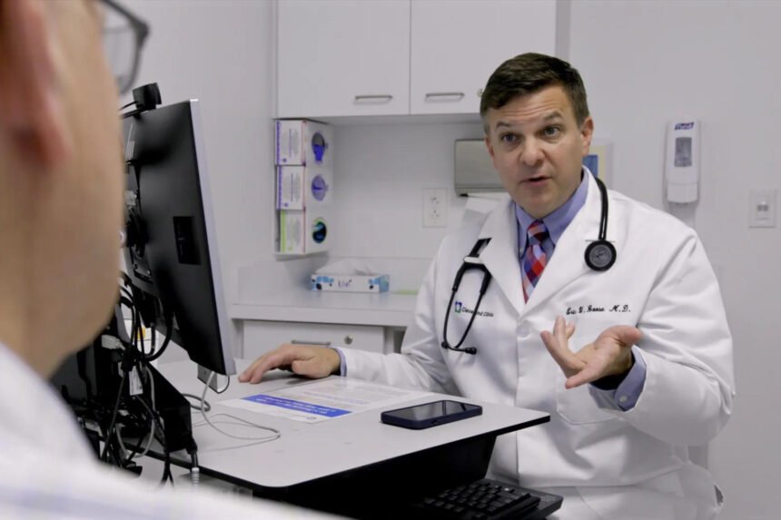 A male doctor in a white coak speaks to someone out of frame.